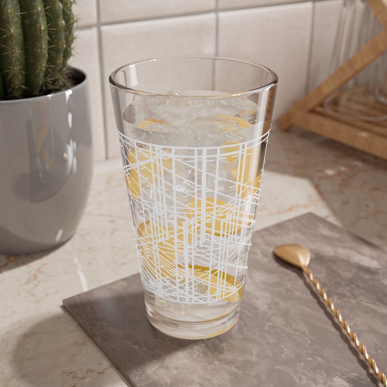 A clear 16oz mixing glass with geometric patterns printed on its surface. It has ice water with lemon slices in it and is sitting on a table.