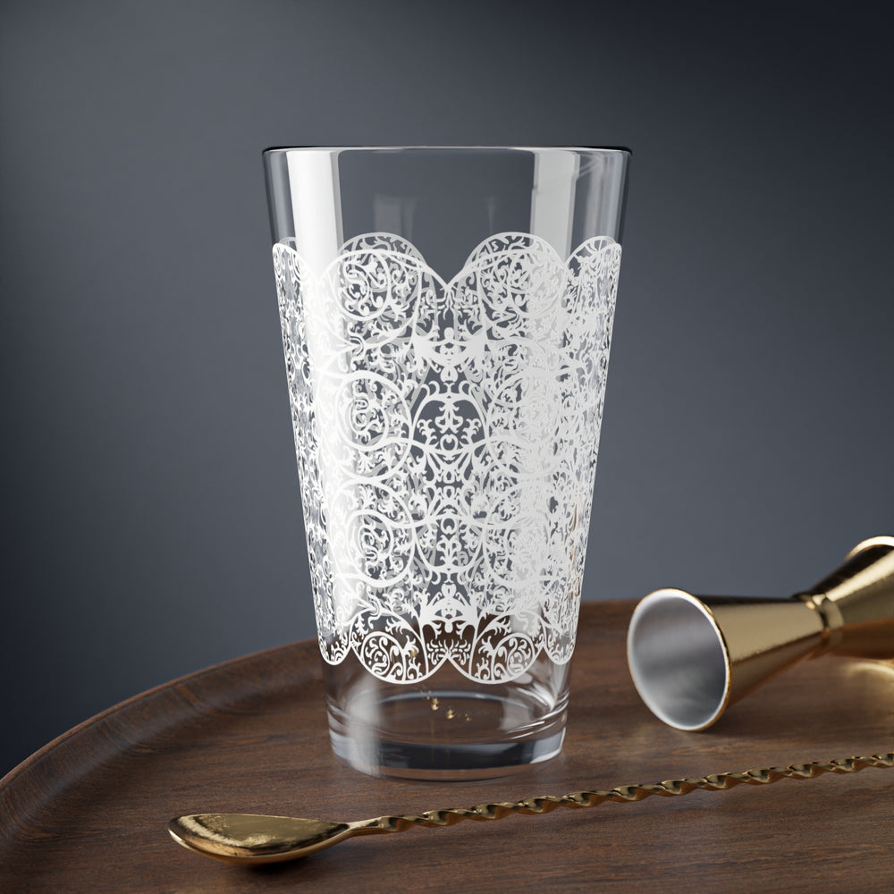 Clear 16 ounce mixing glass with intricate white lace pattern on a wooden tray with gold mixologist drink tools.