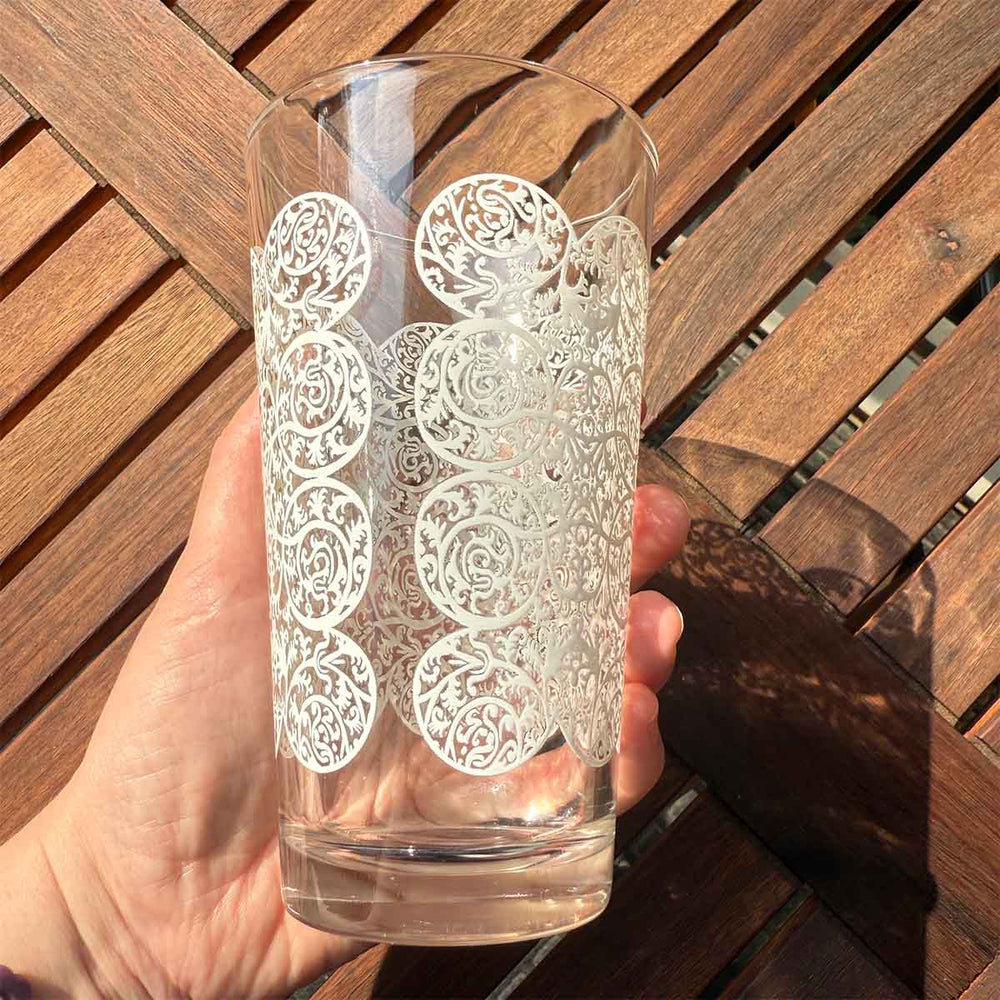 Clear glass with intricate white floral lace patterns held by a hand on a wooden surface