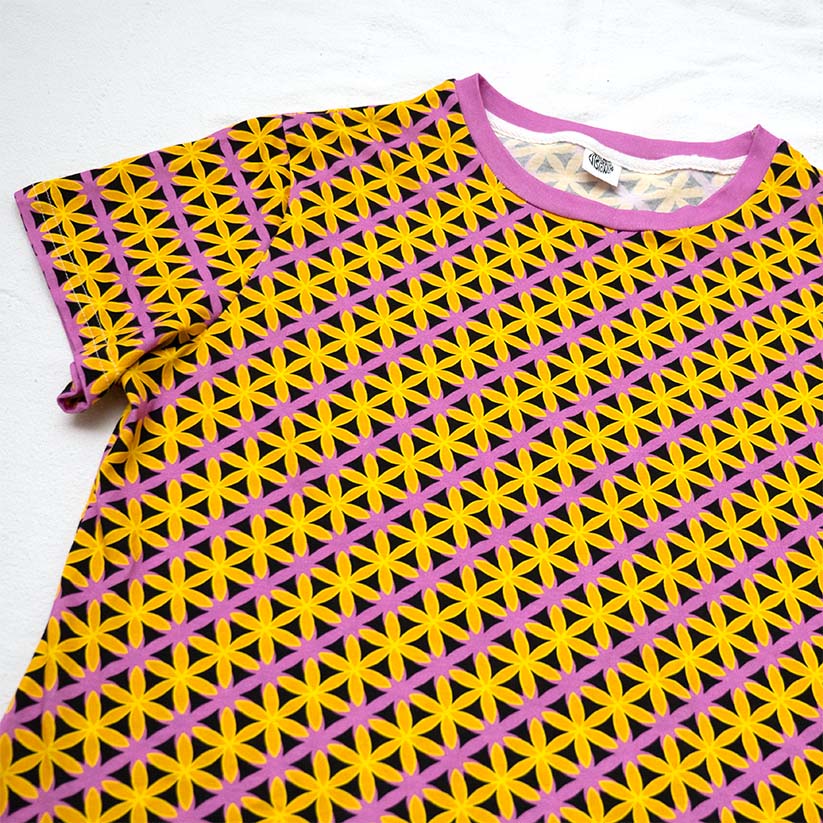 A short-sleeve crewneck t-shirt featuring a vibrant geometric pattern of yellow flowers on a pink grid, with a solid pink collar.