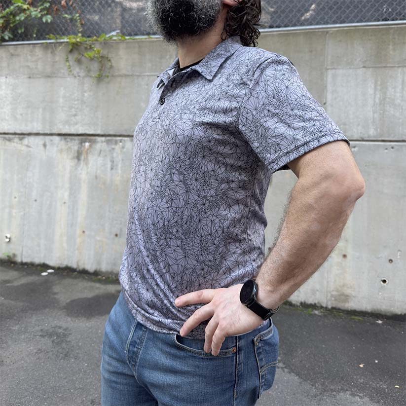 Person wearing Short-sleeve Tri-Mesh Geometric polo  golf shirt with black geometric pattern on a gray background standing in front of a concrete wall 